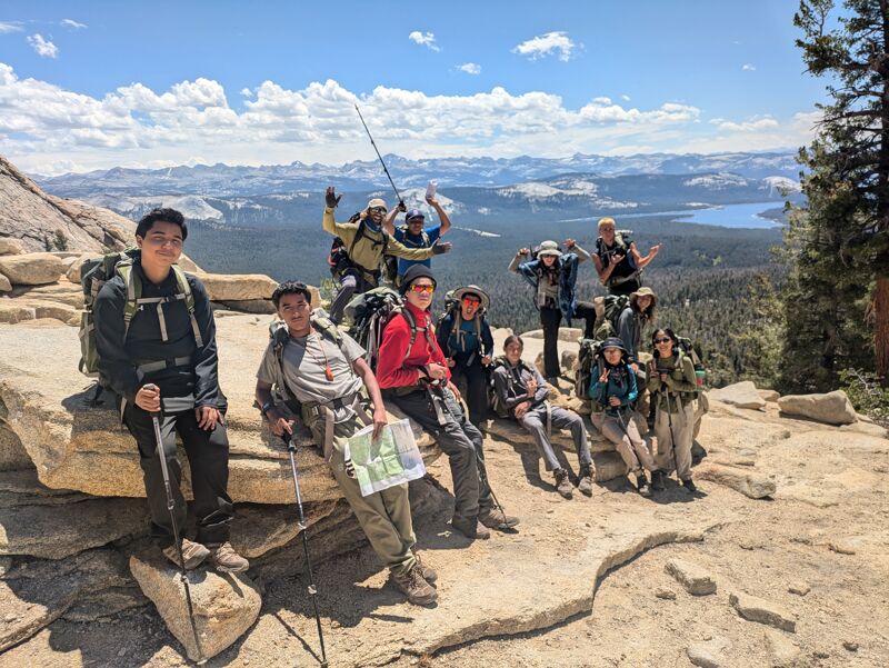 A group of hikers are resting on a rocky outcrop, enjoying a scenic view of a lake and mountains. They are equipped with backpacks and hiking gear, suggesting they are on a trek. The sky is bright with scattered clouds, contributing to the picturesque landscape. The overall atmosphere is one of camaraderie and appreciation for nature.